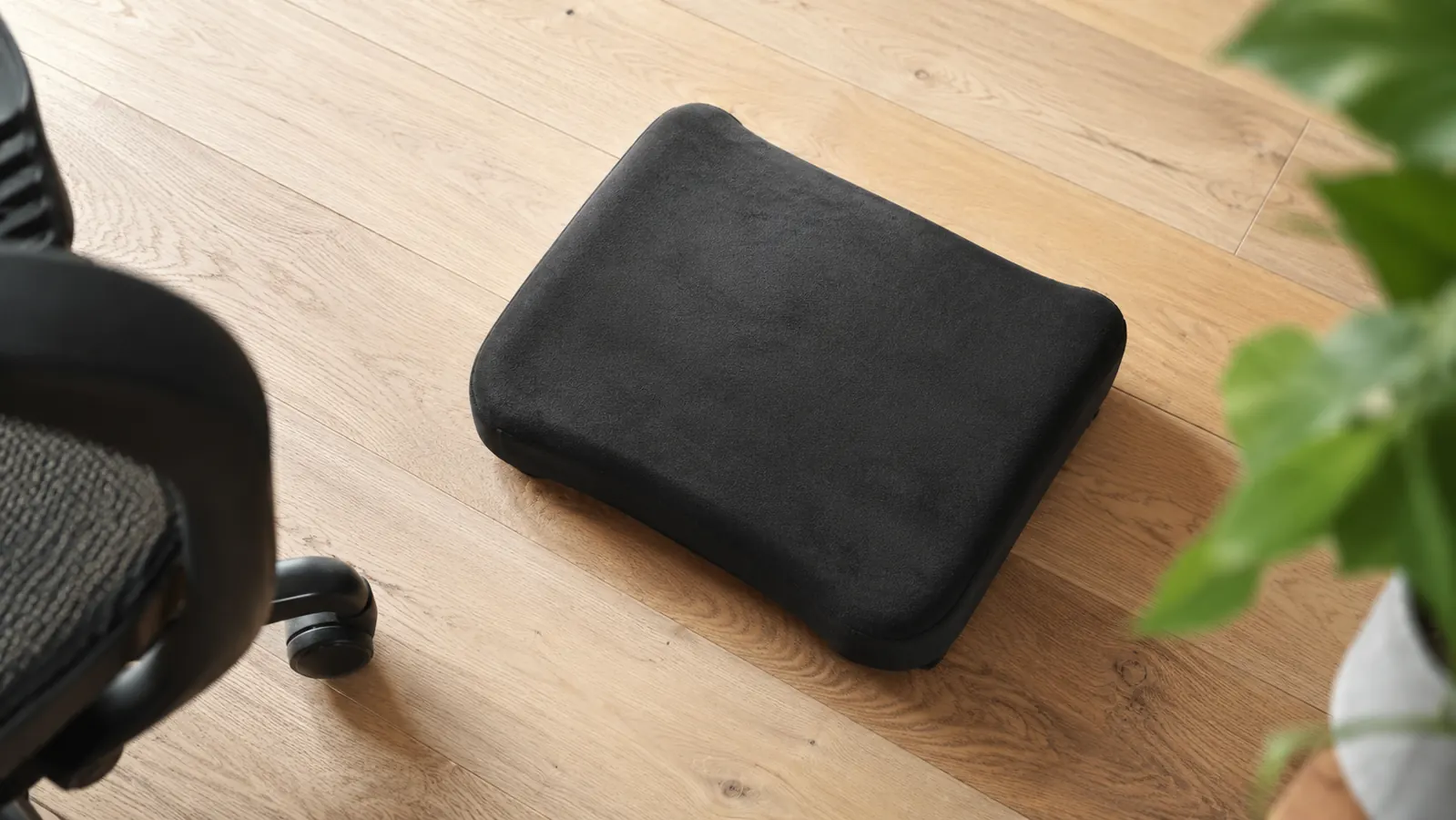 Dark memory foam footrest sitting on light hardwood floor next to a desk chair base, overhead view
