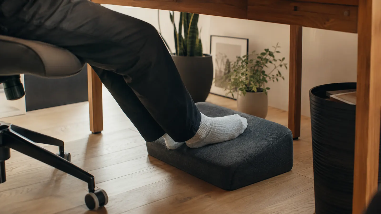 Home office desk setup with soft afternoon light, a person's feet resting on a memory foam footrest beneath a wooden desk