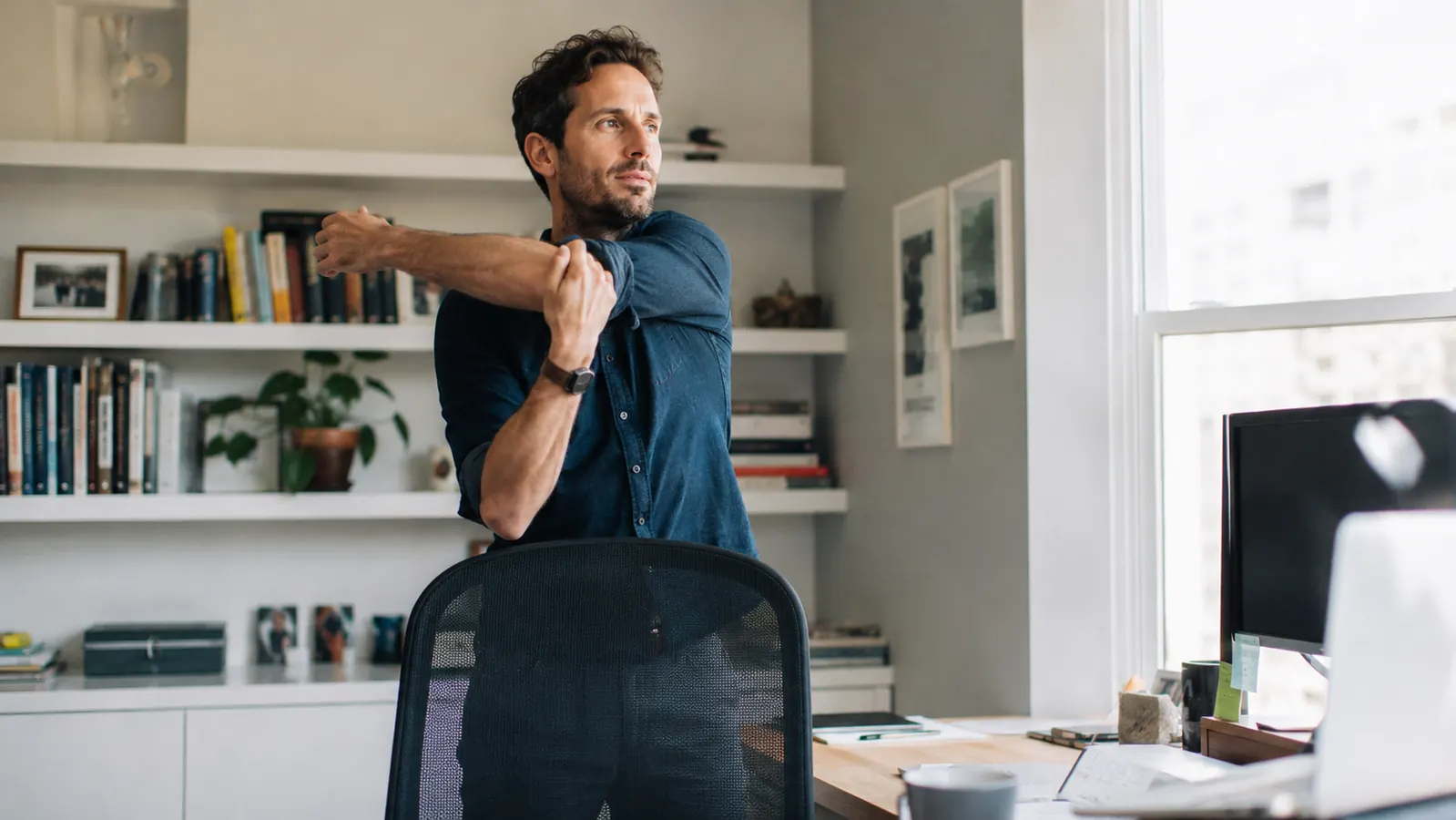 Person standing up from a home office desk chair to take a movement break, stretching their legs in a bright home workspace