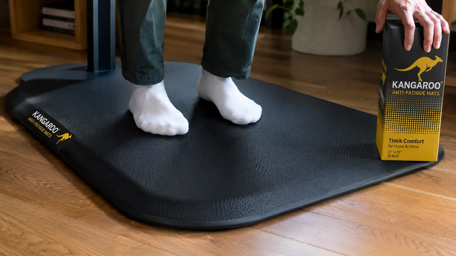 Person standing at a desk wearing socks, feet on a thick black foam mat