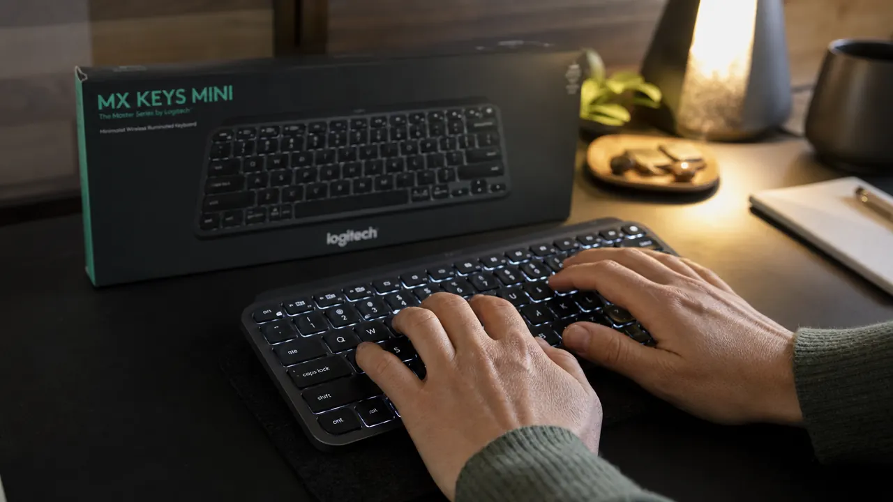 Hands typing on the Logitech MX Keys Mini compact keyboard on a desk next to a monitor